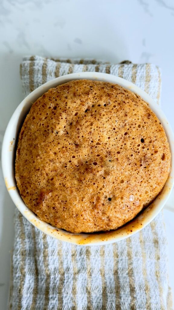 A freshly baked almond butter mug cake in a white ramekin sitting on a striped cloth.