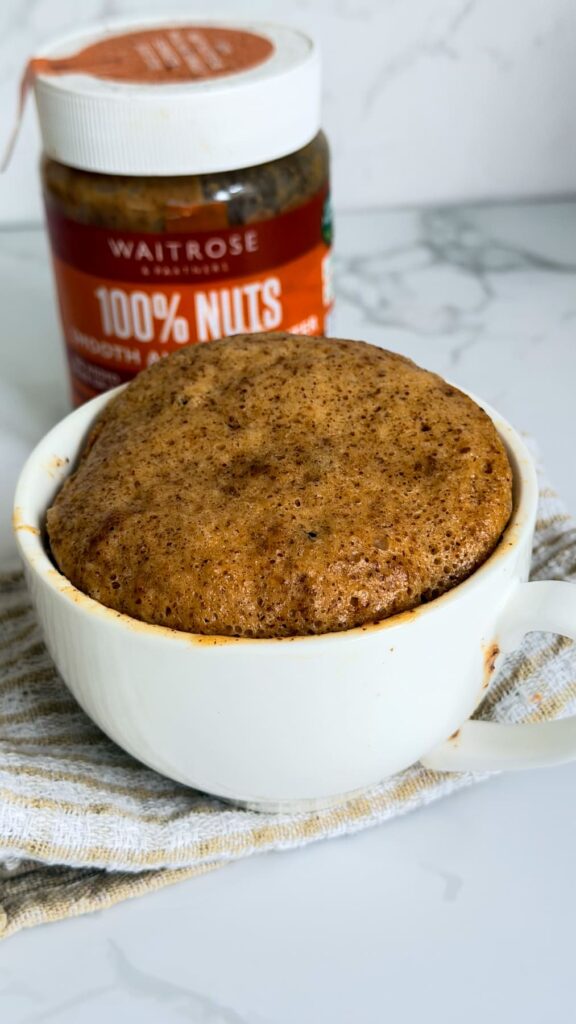 A baked almond butter mug cake next to a jar of Waitrose 100% Nuts smooth peanut butter.