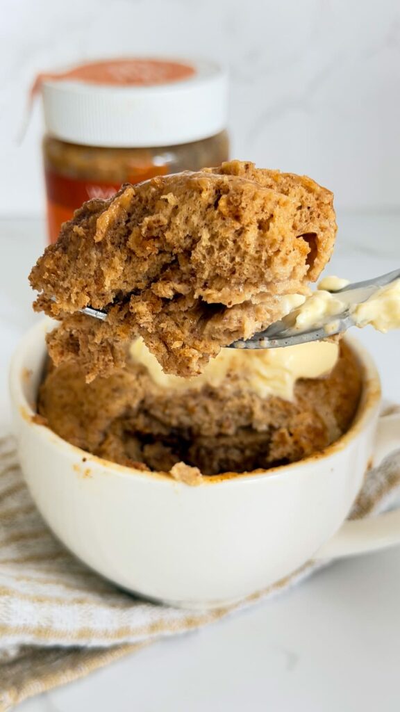 A fork lifts a bite of brown almond butter mug cake topped with a dollop of butter above the remaining cake in a white mug.