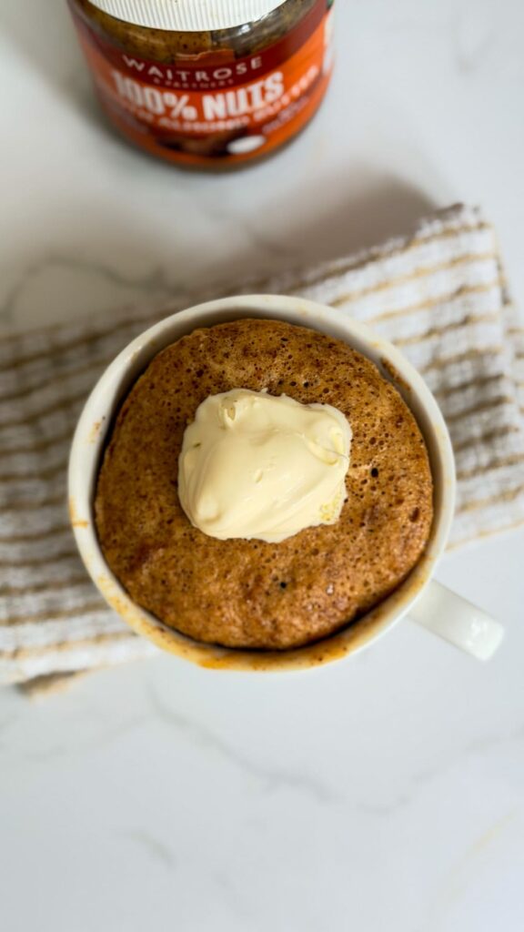 An almond butter mug cake topped with a dollop of butter, with a jar of Waitrose 100% nut butter in the background.