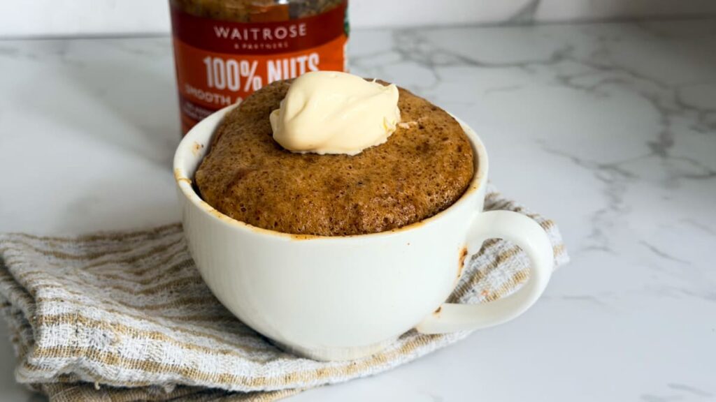 An almond butter mug cake topped with a dollop of cream, with a jar of Waitrose peanut butter in the background.